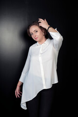 Young beautiful cheerful brunette caucasian adjusting her dark brown hair. Businesswoman with hairstyle in elegant white shirt standing and posing against black wall. Ledy holding hand in hair