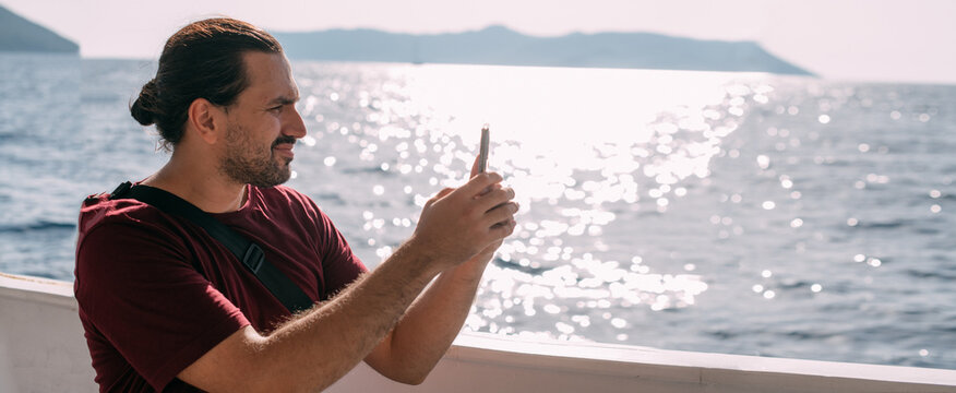 A young, contented man, a tourist rides a boat to the sea, takes pictures on the phone, enjoys a sunny day.