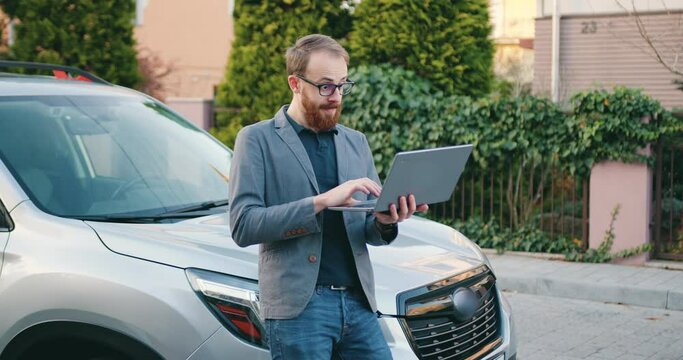Good-looking caucasian business man in jacket using laptop by the car riding good news, outside. Emotional happy man celebrates victory on laptop outdoor.