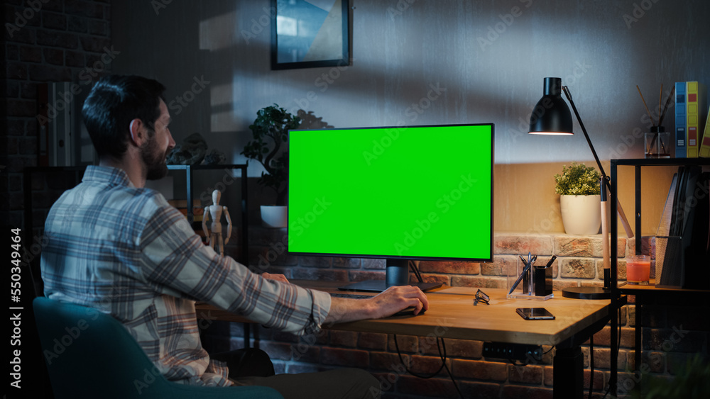 Poster Young Attractive Man Working from Home on Desktop Computer with Green Screen Mock Up Display. Male Checking Corporate Accounts, Messaging Colleagues. Loft Living Room in the Evening