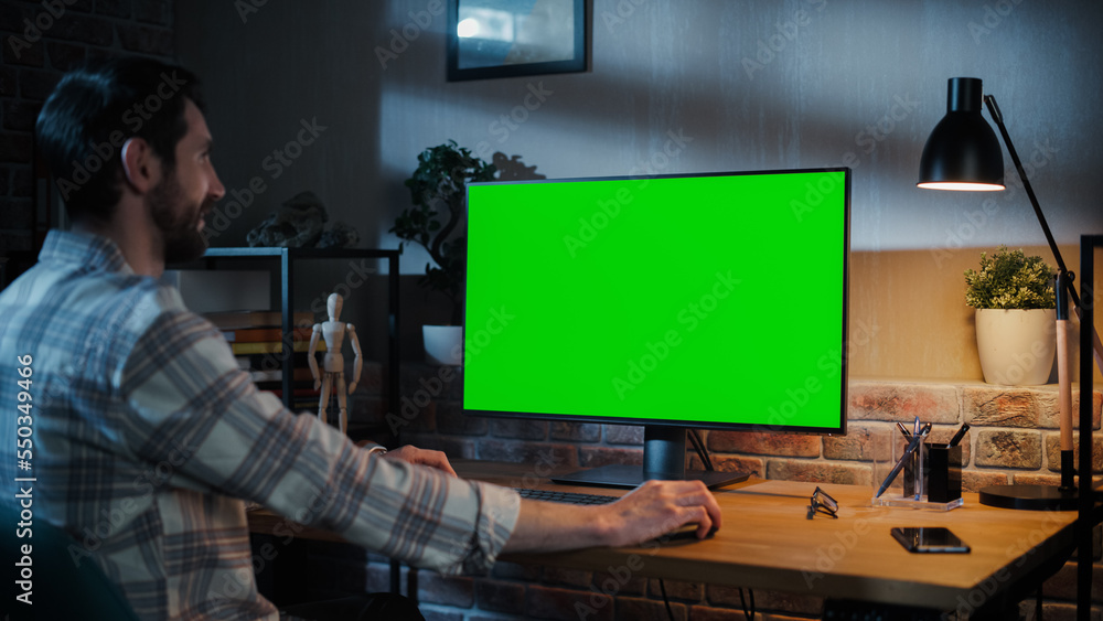 Aufkleber Young Handsome Man Working from Home on Desktop Computer with Green Screen Mock Up Display. Creative Male Checking Social Media, Browsing Internet. Loft Living Room in the Evening