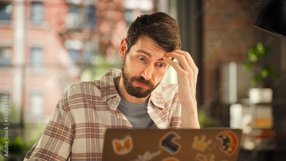 Fototapete Creative Bearded Man Working on Laptop Computer while Sitting at a Table in Stylish Cozy Loft Living Room. Freelancer Working Remotely. Browsing Internet, Using Social Networks, Thinking on Projects.