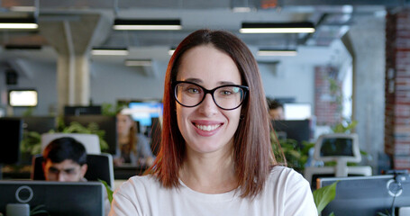 Close up of Caucasian cheerful woman in glasses looking at camera in office. Indoors. Portrait of young pretty female standing in coworking space. Successful leader and businesswoman.