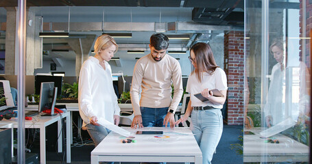 Working team of man and two women standing over the table with tablet device and charts and...