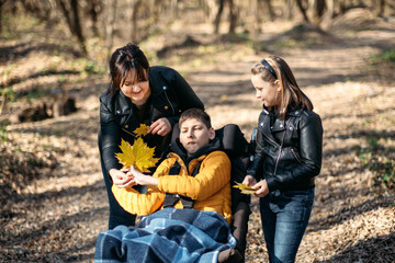 Diversity and inclusion. Happy family, mother, daughter and son teen boy with cerebral palsy spending time together in autumn fall park. Teen boy who uses a wheelchair walking with family