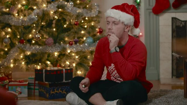 Young man sitting in front of Christmas tree smoking a cannabis joint in a red Santa hat