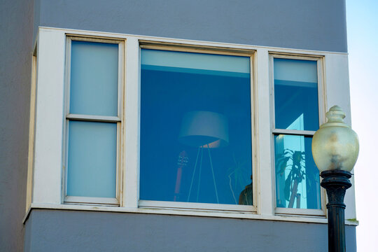 Hidden lamp visible through open window with no curtains with white accent paint and gray stucco facade on house in shade