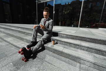 positive businessman in roller skates chatting on smartphone while sitting near paper cup on stairs.