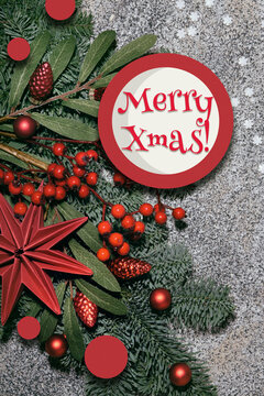 Merry Xmas text, greeting on round Christmas paper card. Flat lay with fir twigs decorated with red rowan berry, paper star and glass trinkets on grey textured stone background.