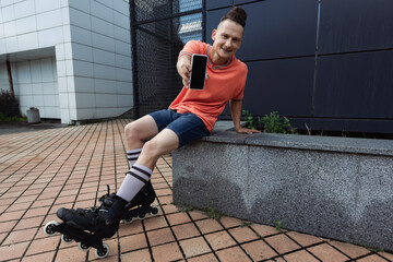 Smiling man in rollers holding smartphone with blank screen on urban street.