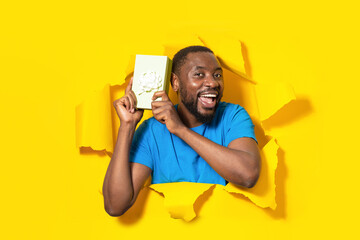Happy black man holding gift box and smiling at camera, posing in hole of yellow paper background, collage, copy space
