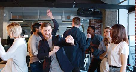 Young men and women dancing and having fun in office space. Friday after work. Multiethnic team of coworkers having party at work. Cheerful Caucasian man boss with employees. - obrazy, fototapety, plakaty