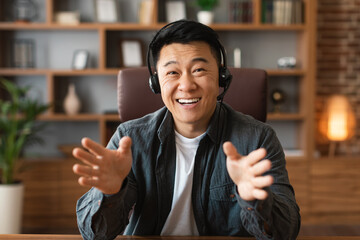 Cheerful adult japanese male with headphones gesticulates hands, looks at computer webcam
