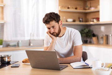 Boring tired sad adult caucasian man with beard in white t-shirt watch online lesson at laptop in modern kitchen