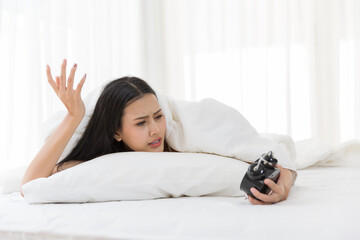 Asian young woman hates waking up early in the morning. Sleepy girl looking at alarm clock with upset feeling on bed in the morning at home