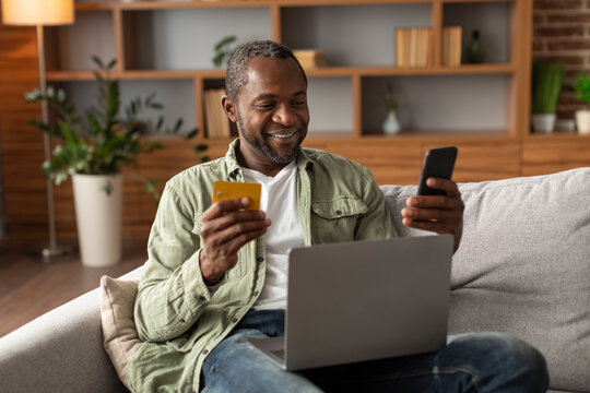 Cheerful middle aged african american man sit on sofa with laptop uses credit card and smartphone for online shopping