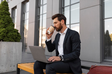 Happy busy young european man manager with beard in suit typing on computer, drink cup of coffee takeaway