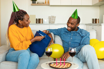 Happy birthday my love. Excited black woman giving gift to her husband, young couple sitting on...