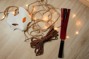 Shibari ropes and flogger and white mask before a session over a wooden floor
