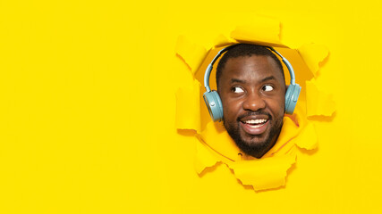 Black man wearing headphones, listening to music and looking aside at free space through hole in torn yellow paper