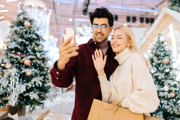 Portrait of friendly young couple talking looking at smartphone having video call, giving online New Year greetings standing in hall of celebrate shopping mall with xmas decorations in Christmas eve
