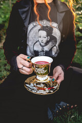 Red haired girl with dark clothes (and no head) drinking a red drink ('blood') in a old centuries tea cup and saucer in the park