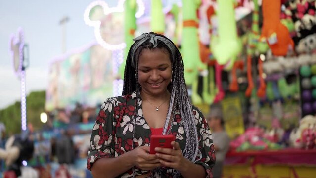 African American young woman with braided hair at amusement park having fun and using mobile to take funny selfies.