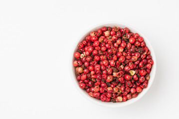 Fragrant spice pink pepper seeds in a small white ceramic bowl with copy space.  Pink peppercorn isolated top view with copy space