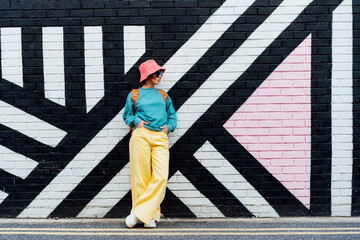 Hipster fashion young woman in bright clothes, sun glasses and bucket hat posing on the painted brick wall background. Urban city street fashion. Selective focus. Copy space