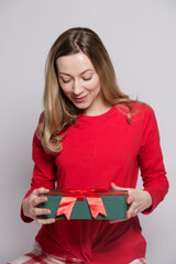 White european young woman with blonde hair in red New Year's pajamas with a gift in her hands looks at the gift isolated on gray background