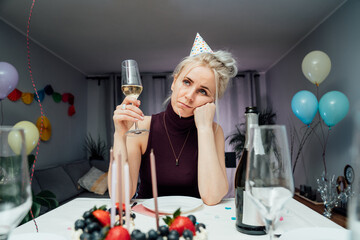 Bored, sad attractive woman drinking champagne while celebrating birthday at home, sitting alone at...
