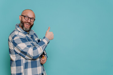 Excited caucasian mid adult beardy male in plaid shirt and glasses gestures thumb up toothy smiles against turquoise studio backdrop looks at camera. Mockup, sale, discount, promo. Teacher with book.
