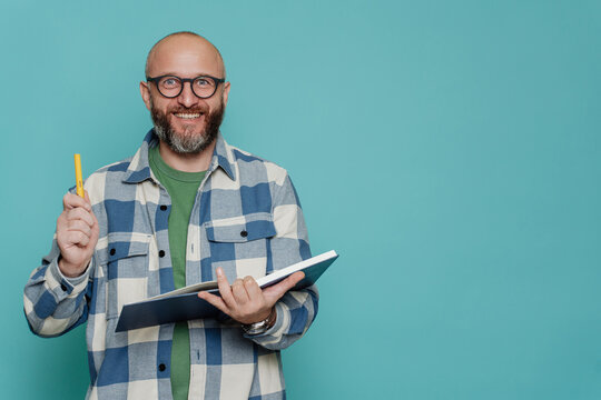 Cheerful Caucasian bald beardy man in plaid shirt holds book and pen looks at camera against turquoise  studio backdrop. Excited Italian male in glasses, teacher has idea. Mockup, education, knowledge