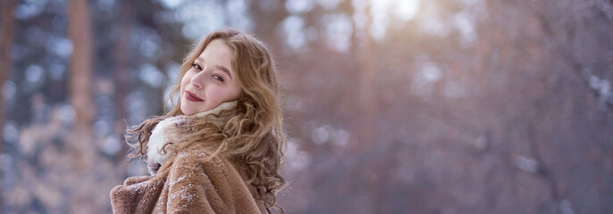 A white blonde girl in a knitted hat, a long scarf and a fur coat walks through the forest and smiles. Woman catches snow in white mittens. Winter is on the street. High quality photo. Christmas Eve