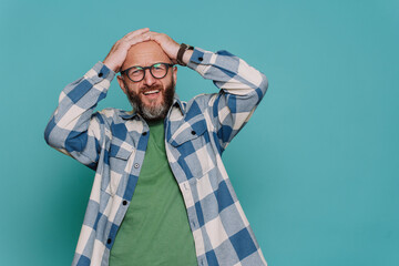 Disappointed mid adult caucasian beardy man in plaid casual shirt and spectacles holds head in frustration looks at camera with upset face expression against turquoise studio background. Mockup