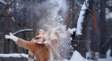 A white blonde girl in a knitted hat, a long scarf and a fur coat walks through the forest and smiles. Woman catches snow in white mittens. Winter is on the street. High quality photo. Christmas Eve
