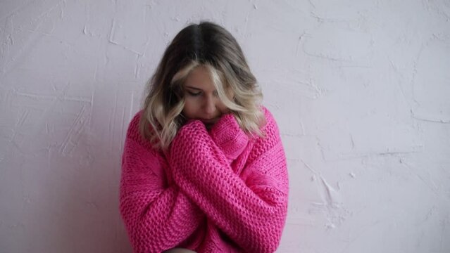 Woman in home room without central heating sitting near white wall. Young girl in warm pink pullover get warm. Concept of problems with central heating. Flu symptoms, disease, chill, depression