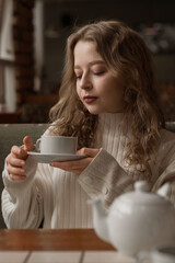 White blonde girl in a warm sweater drinks tea in a coffee shop. In a cafe, coffee is poured into a white mug. Hot tea for morning breakfast. Warming coffee in the restaurant on a winter evening. High