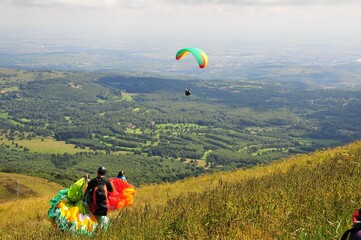 Person paragliding above a scenic green landscape and another one walking with the parachutes