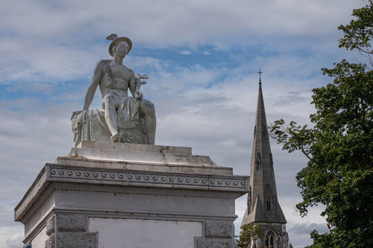Copenhagen, Denmark - July 24, 2022: Mercurius or Hermes God white stone statue on pillar at east side entrance to Churchill Parken. Blue cloudscape, St. Albans church spire, green foliage