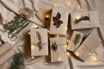 Many Christmas gift boxes, eco-friendly wrapping style. Craft brown paper with trendy folds and natural twigs. Lights, classic scissors, linen textile