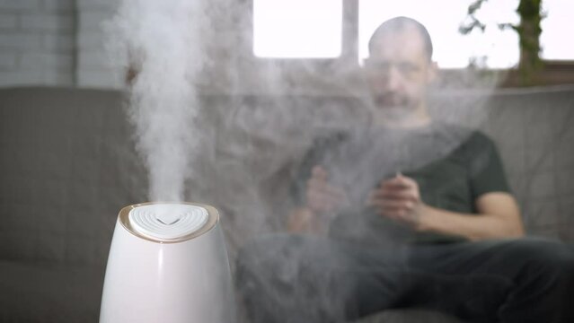 Home humidifier. Man by air humidifier in room. A male sits with his ipad on the sofa and enjoy the air from purifier.