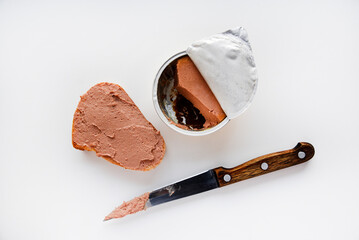 Meat pate in a jar on a white background. Spreading pate on bread with a knife. Meat pate, knife and bread. Delicious breakfast of pate.