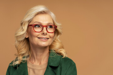 Portrait of smiling mature woman wearing red stylish eyeglasses looking away isolated on beige background, copy space. Vision concept 