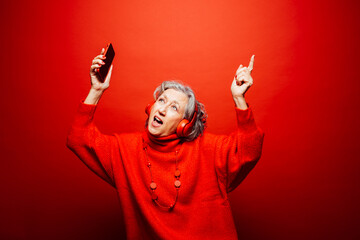 Senior woman wearing red clothes, with red headphones and red smartphone, dancing over a red background