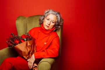 Portrait of a senior woman wearing red clothes and sitting on a green armchair with a bouquet of red roses, over a red background