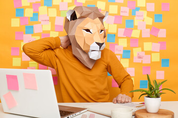 Indoor shot of sick tired unknown man in lion mask wearing orange jumper sitting at workplace with...