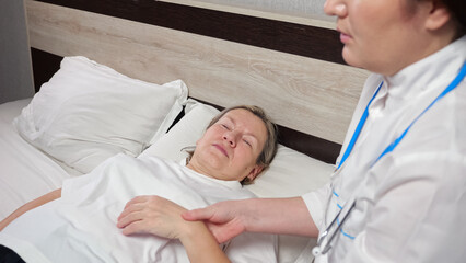 Experienced therapist with stethoscope does examination of woman lying in bed. Family doctor checks pulse and temperature of sick patient at home closeup
