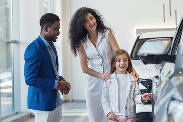 Mother and daughter choose together nice car with African American male dealer help in showroom....