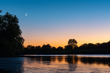 Sunset over a lake surrounded by trees and a crescent moon